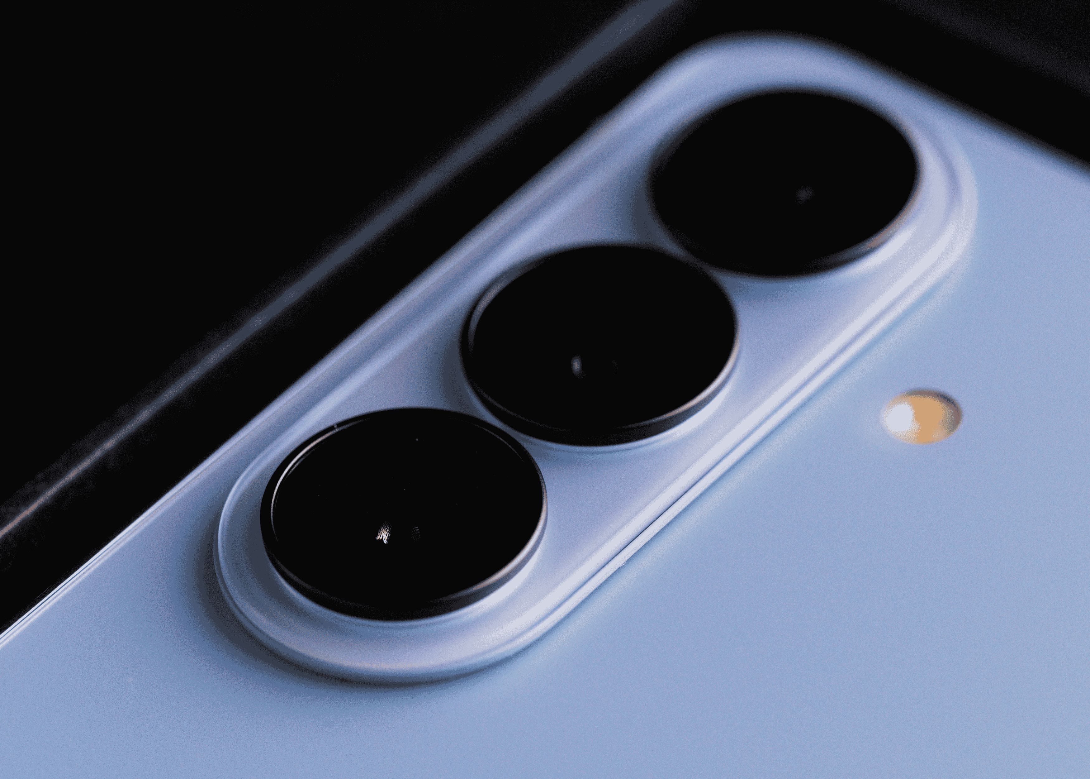A close-up shot of the back of a light blue smartphone, showing a triple camera array with black lenses and a small flash to the right.